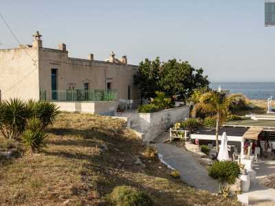Sulla spiaggia di Porto Ghiacciolo, a Monopoli, tra bar e ombrelloni, si affaccia una masseria in pietra chiara e dalle linee squadrate, colorata da porte in legno azzurre e ringhiere in ferro dipinte di verde
